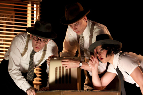 A production photo from HardBoiled. The cast are all leaning into the middle to listen to an old radio.