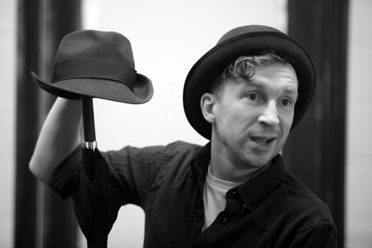 A black and white photo of Jeremy Legat. He is a holding a hat.