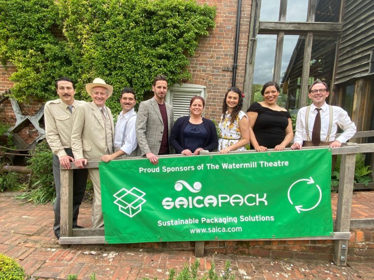 The cast of Our Man In Havana and staff from Saica Pack standing outside The Watermill Theatre with a large green banner with Saica Pack's credentials.